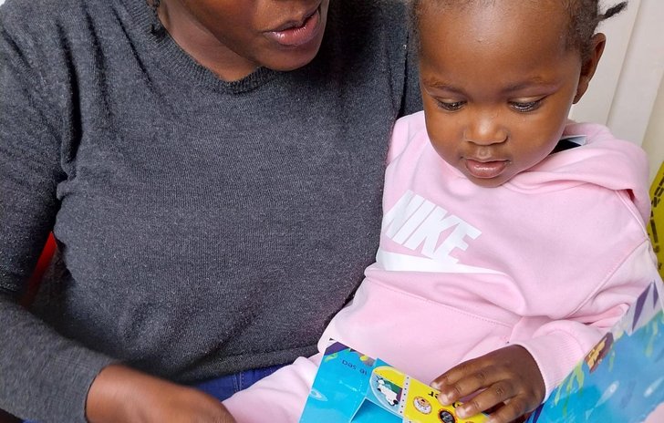An adult and a child are sitting together, reading a colorful children's book. The adult is wearing a dark gray sweater and blue jeans with ripped knees, while the child is dressed in a light pink hoodie and matching trousers..
