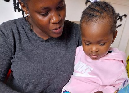 An adult and a child are sitting together, reading a colorful children's book. The adult is wearing a dark gray sweater and blue jeans with ripped knees, while the child is dressed in a light pink hoodie and matching pants.