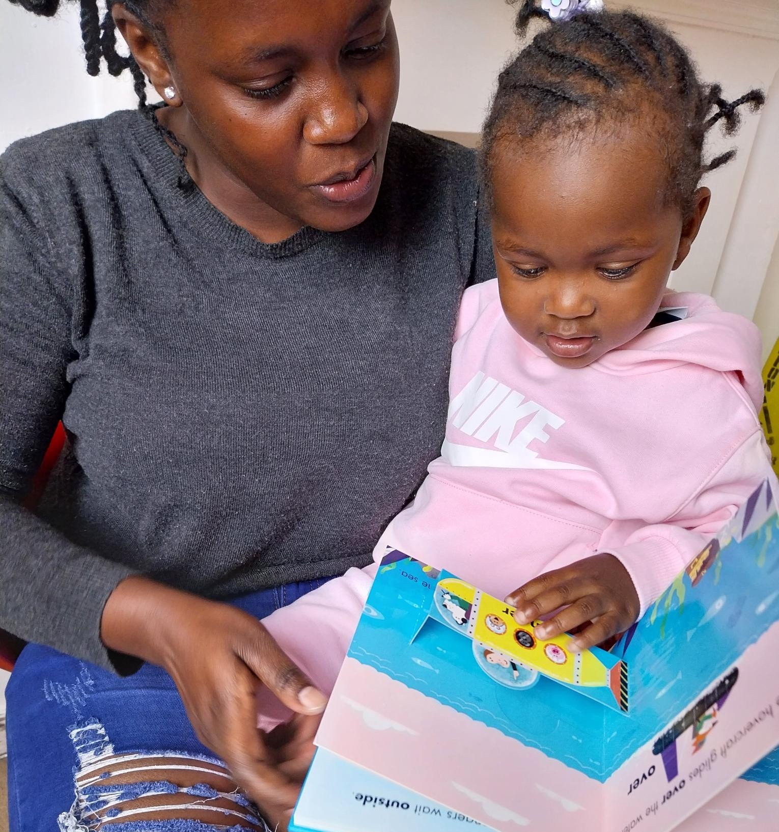 An adult and a child are sitting together, reading a colorful children's book. The adult is wearing a dark gray sweater and blue jeans with ripped knees, while the child is dressed in a light pink hoodie and matching pants.
